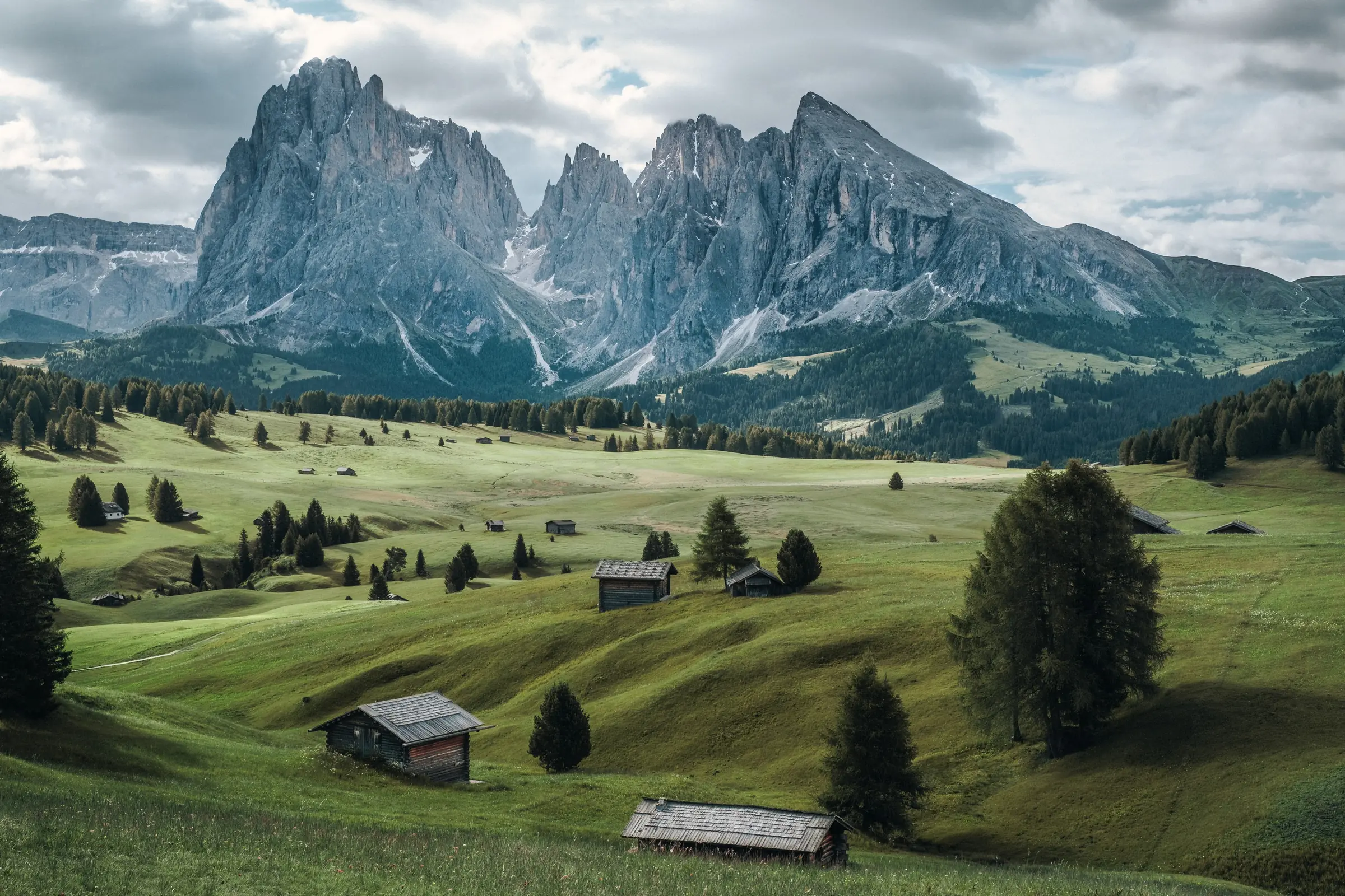 Wooden huts scattered across the green meadows of Seiser Alm, below the Sassolungo and Sassopiatto peaks in the Dolomites